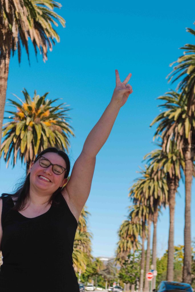 woman closing her eyes and smiling with a peace sign on her outstretched arm with a street lined with palm trees and the Hollywood Sign behind her