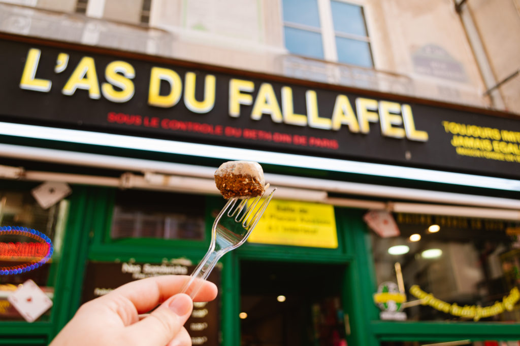 Fallafel on a fork being held up by a hand from the left with L'as du fallafel restaurant in the background
